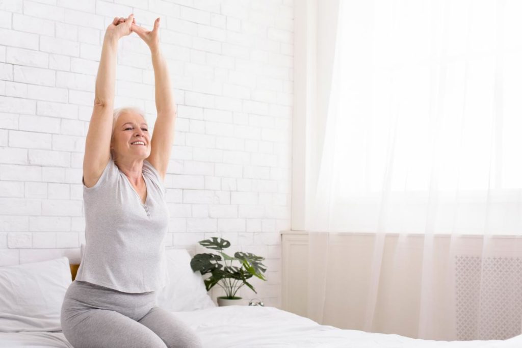 older woman sitting on her bed in sunlit bedroom performing useful exercises for seniors