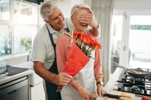 senior man has snuck up behind his senior wife and covered her eyes with one hand while holding bouquet of roses with other hand all as part of valentines day activities for seniors