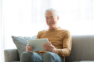 male senior citizen seated on couch and using a tablet computer as he experiences the impact of technology on senior care in skilled nursing centers